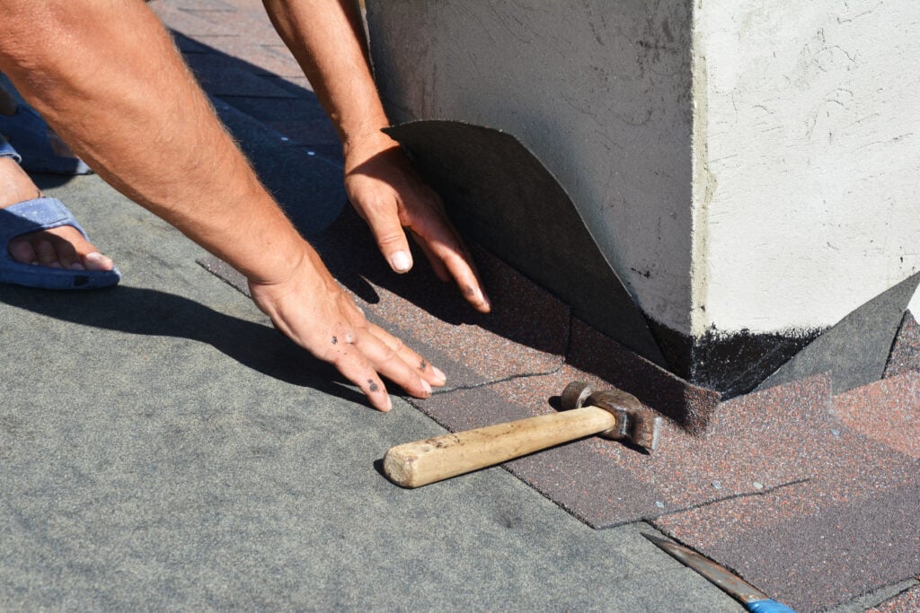 storm damage roof insurance claim Asphalt shingles roofing construction. Roofer repair and laying asphalt shingles with vapor barrier in chimney problem area. Chimney waterproofing on house rooftop.