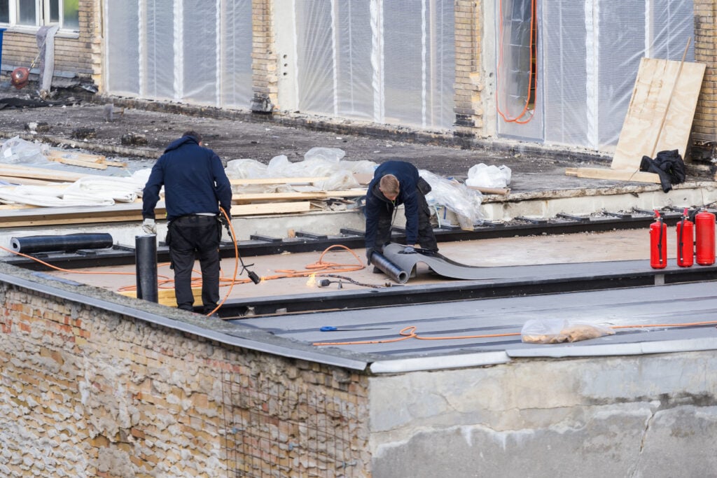 Two construction workers install a black waterproofing membrane and asphalt bitumen shingle on a flat roof, while another worker stands nearby on top of the building