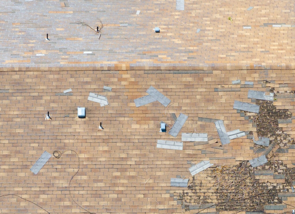 Aerial view reveals a damaged roof. Shingles blown off during a storm and scattered. Structure requires urgent repair. Ideal for insurance or construction contexts