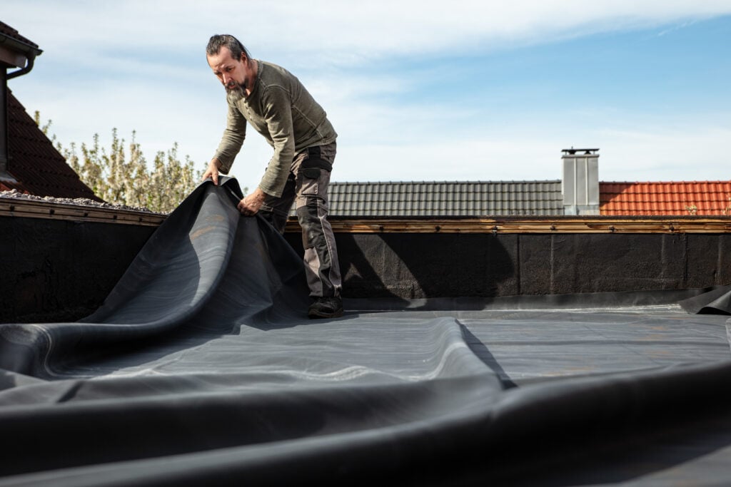epdm roofing A craftsman rolls out a thick rubber tarpaulin on a flat roof to re-seal and repair the broken bitumen coating